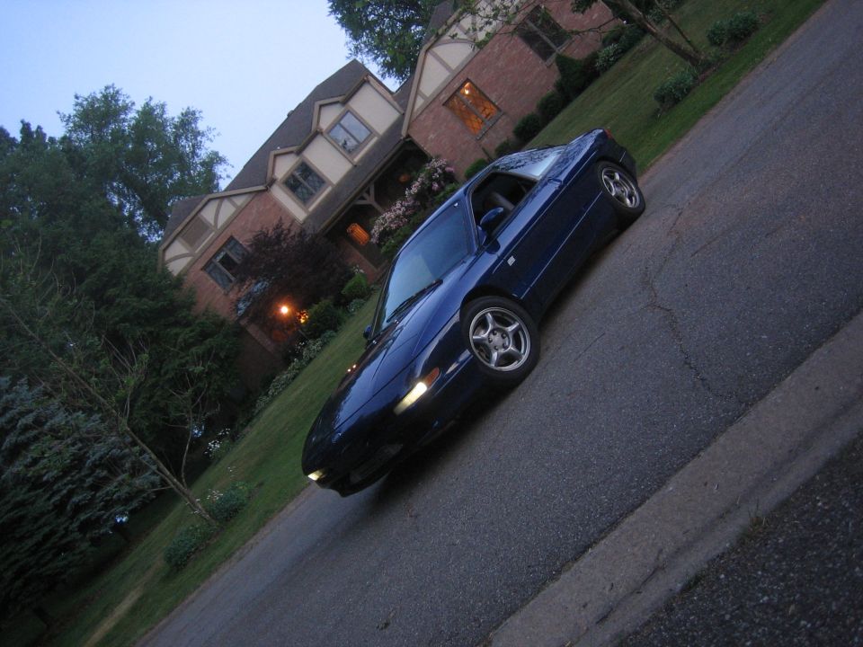 Blue coupe parked on a street in front of a house. The car is angled towards the viewer