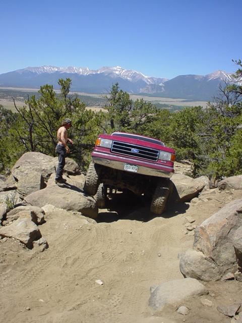1991 Ford Bronco - A red 1991 Ford Bronco tackles a challenging rocky off-road path, with a scenic mountain range visible in the distance.