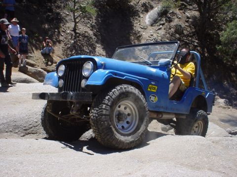 1991 Ford Bronco - A blue Jeep CJ navigates a rough, rocky terrain with spectators watching from a hillside.