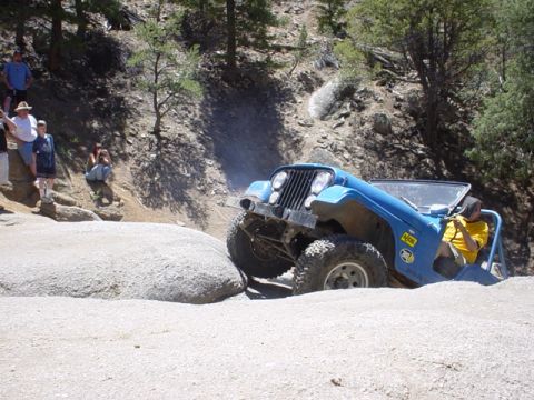 1991 Ford Bronco - A blue Jeep Wrangler maneuvers over rocks on an off-road trail, with spectators watching from a hillside.