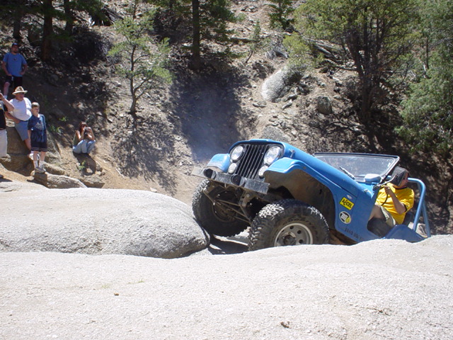 Blue Jeep Wrangler navigating a rocky off-road trail, front right tire on a large boulder