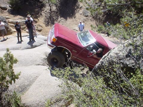 1991 Ford Bronco - A red Ford Bronco is shown off-roading on a rocky terrain with several people observing.