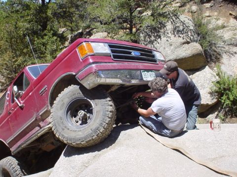 1991 Ford Bronco - Two people work on the undercarriage of a maroon 1991 Ford Bronco that is stuck on rocks.