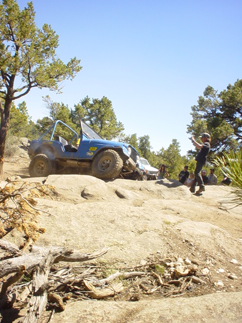 1991 Ford Bronco - A blue off-road vehicle navigates a rocky trail under a clear blue sky, with spectators observing the action.