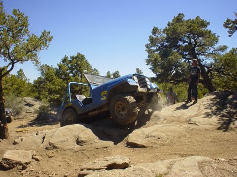 1991 Ford Bronco - A blue Jeep CJ-7 navigates a rocky trail, kicking up dust.