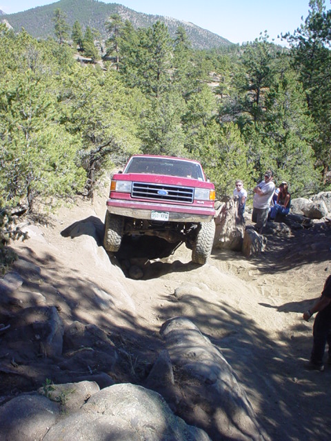 1991 Ford Bronco - Red 1991 Ford Bronco tackling a challenging off-road course in a mountainous, wooded area.