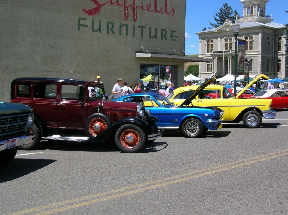 Blue Ford Mustang coupe with hood up at outdoor car show beside maroon vintage sedan and yellow classic car