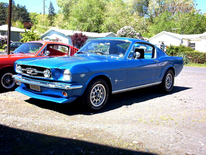 Blue 1965 Ford Mustang fastback on gravel driveway, chrome bumpers and styled steel wheels