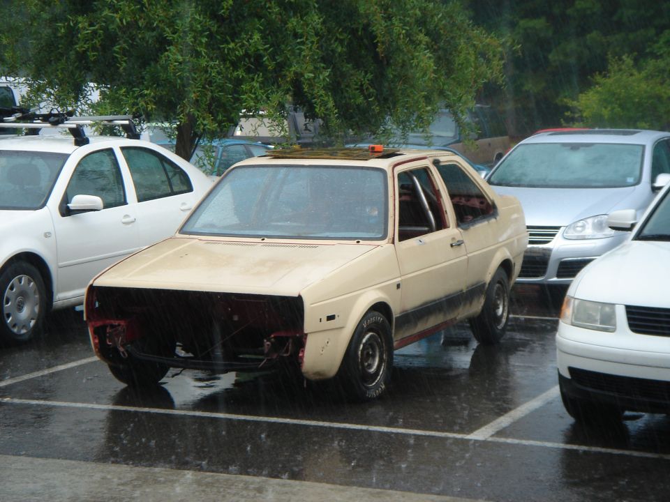 Beige Volkswagen Jetta, missing front bumper and grille, parked in rain. Roll cage visible inside. Other cars nearby