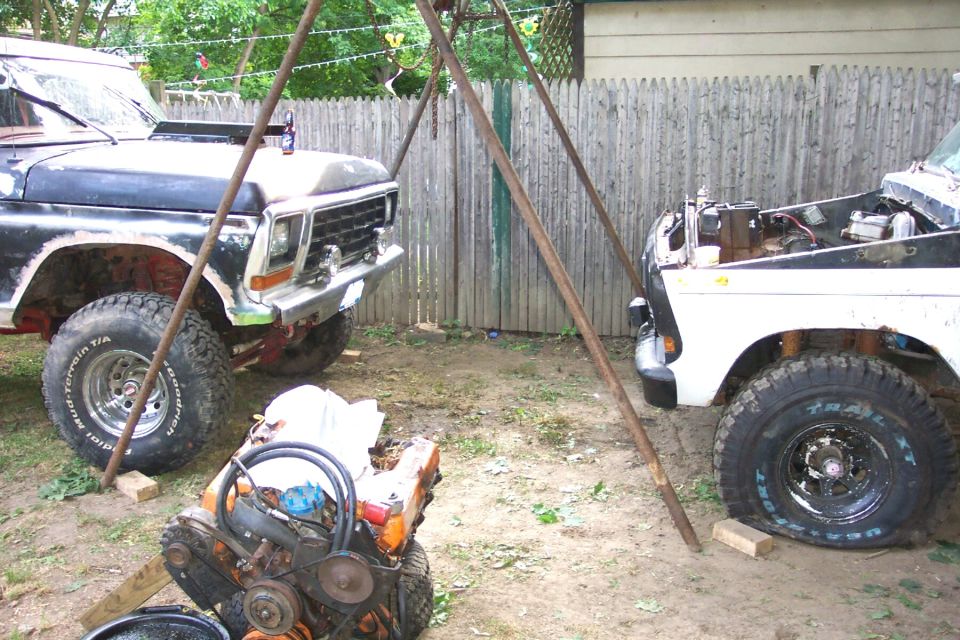 Two project Ford 4x4 trucks by a fence with a V8 engine on the ground