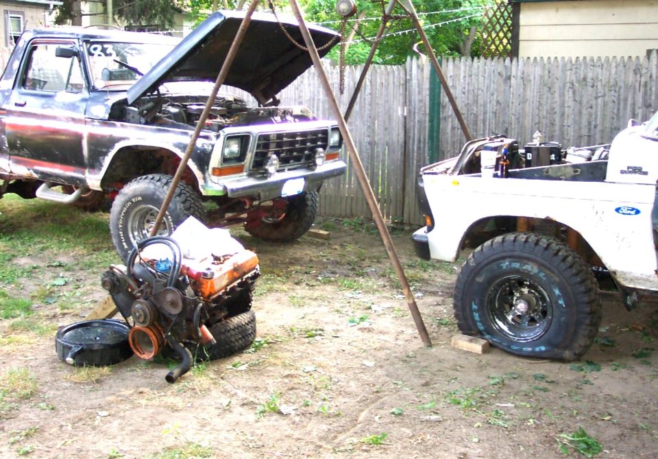 Two open-hood Ford trucks in a yard with an engine on the ground under a hoist
