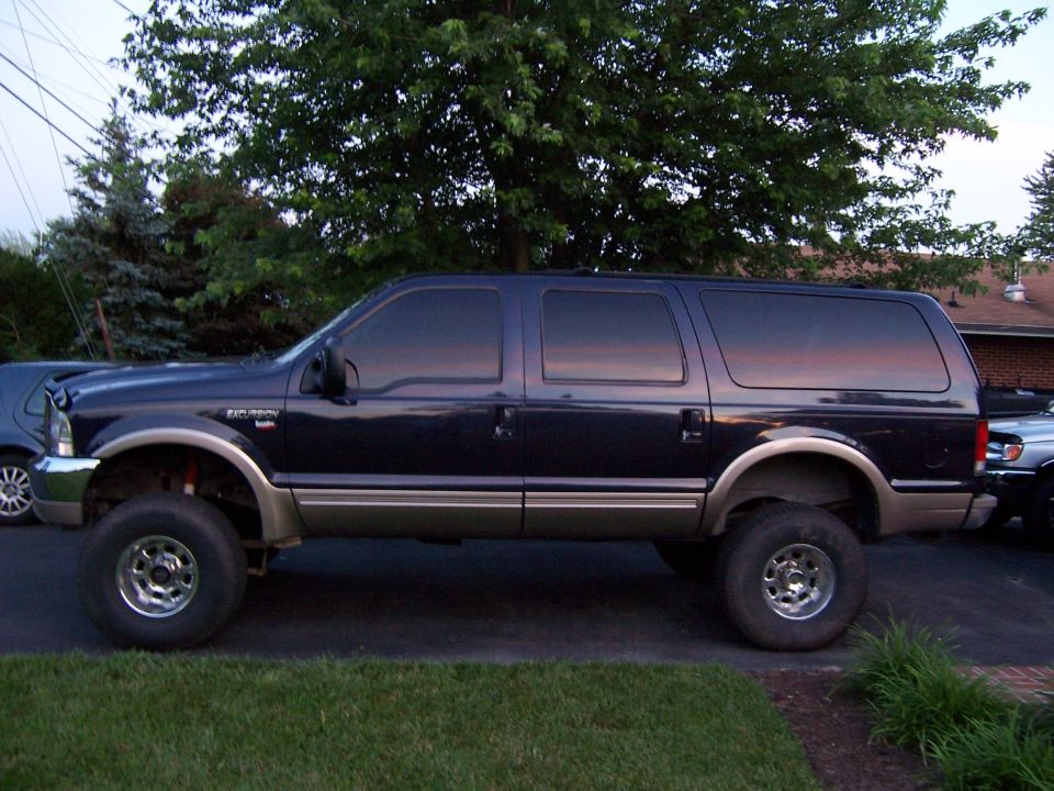 Lifted dark blue Ford Excursion with tan lower cladding on chrome wheels in a yard