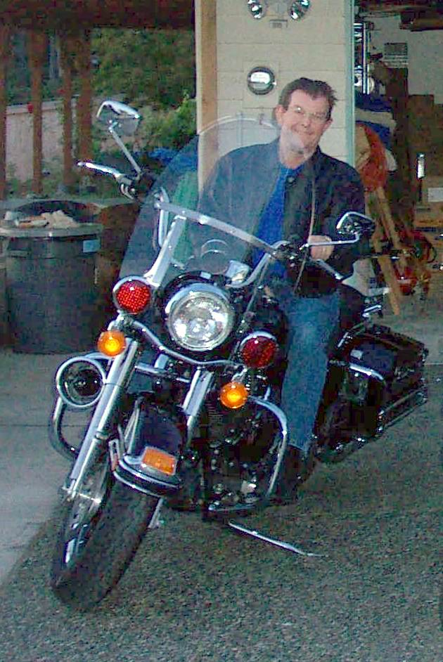 Man seated on a black Harley-Davidson touring motorcycle with windshield and crash bars in a garage