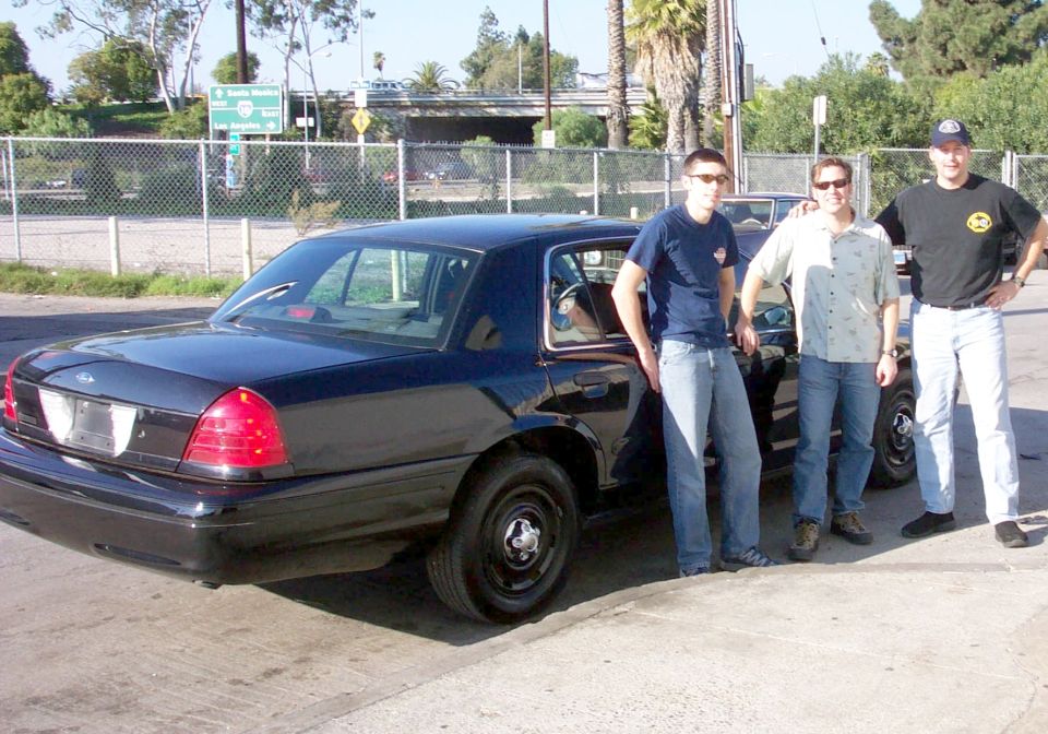 Dark 2003 Ford Crown Victoria, rear quarter view, with three men standing beside it