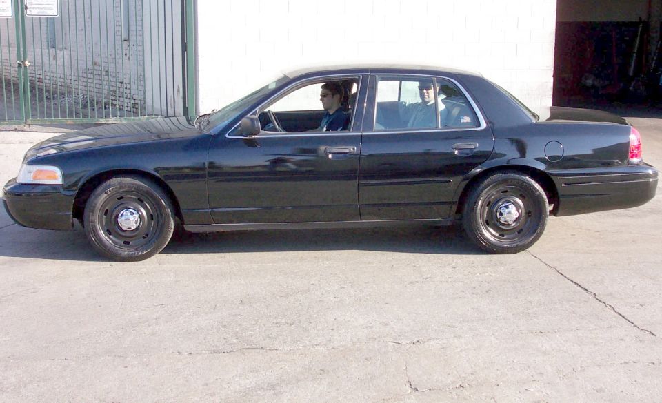 Black 2003 Ford Crown Victoria sedan parked on concrete, side view. Two people inside