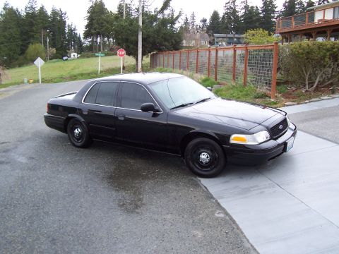 2003 Ford Crown Victoria - A black sedan is parked on a wet road next to a sidewalk, with a residential area in the background.