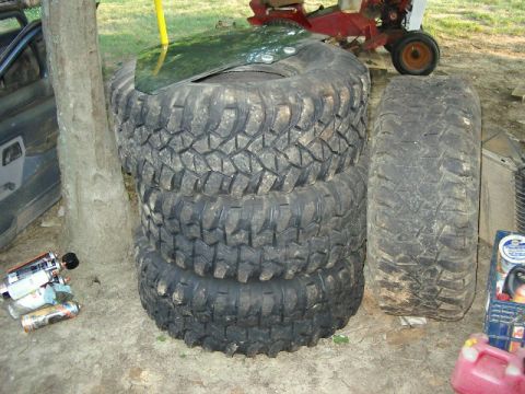 1985 Ford Bronco - Four large off-road tires are stacked, with a windshield placed on top. A vehicle door is visible to the left.
