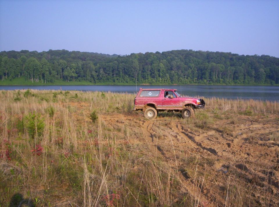 Red Ford Bronco with mud splatters driving on a dirt hill near a lake