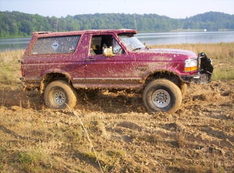 1995 Ford Bronco - This 1995 Ford Bronco is covered in mud as it navigates a challenging off-road terrain by a lake.