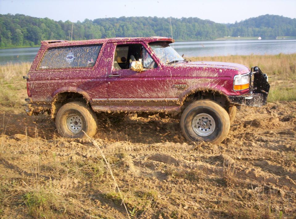 Mud-covered red 1995 Ford Bronco driving through mud near a lake