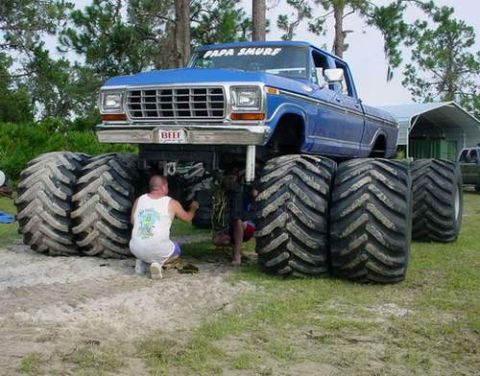 1990 Ford Bronco - blue monster truck lettered 'Papa Smurf' rides on four pairs of giant tires while a man kneels working beneath it on sandy ground.