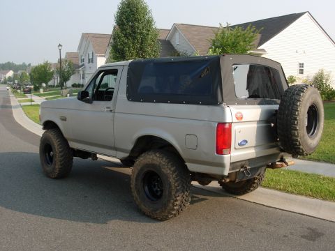 1995 Ford Bronco - Silver Ford Bronco with a black soft top and spare tire on the rear is parked on a suburban street.