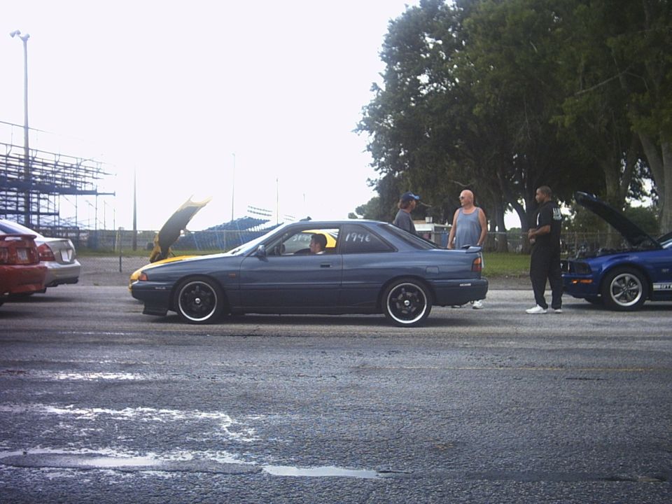 Blue-gray coupe with yellow hood open and '1946' on window at a drag strip, people and a blue Mustang nearby