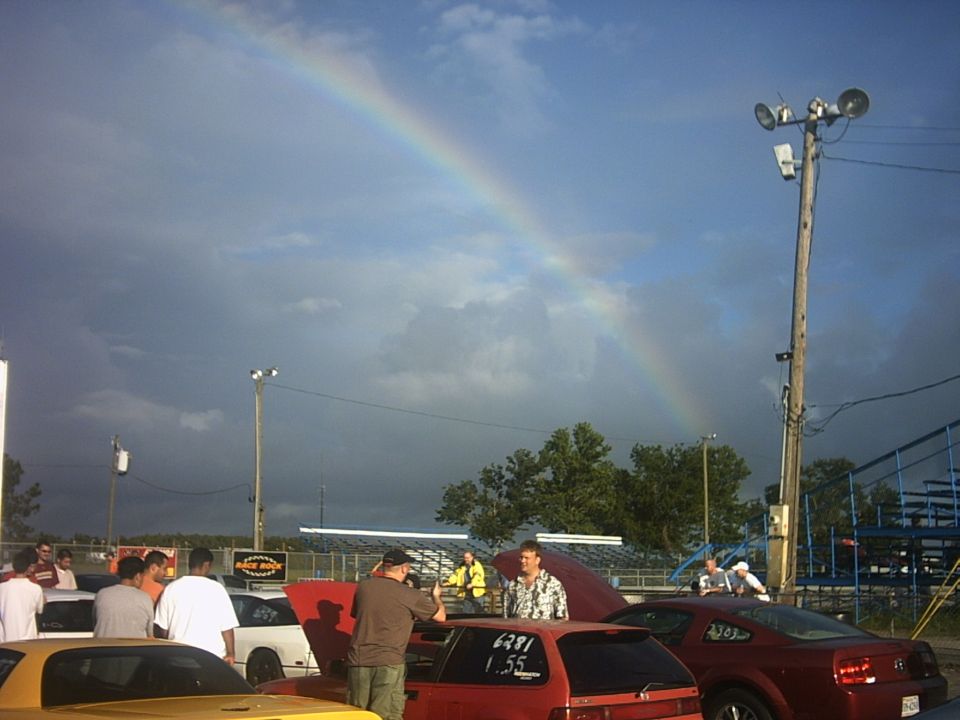Rainbow over a dirt-track racing pit area with spectators and parked race cars