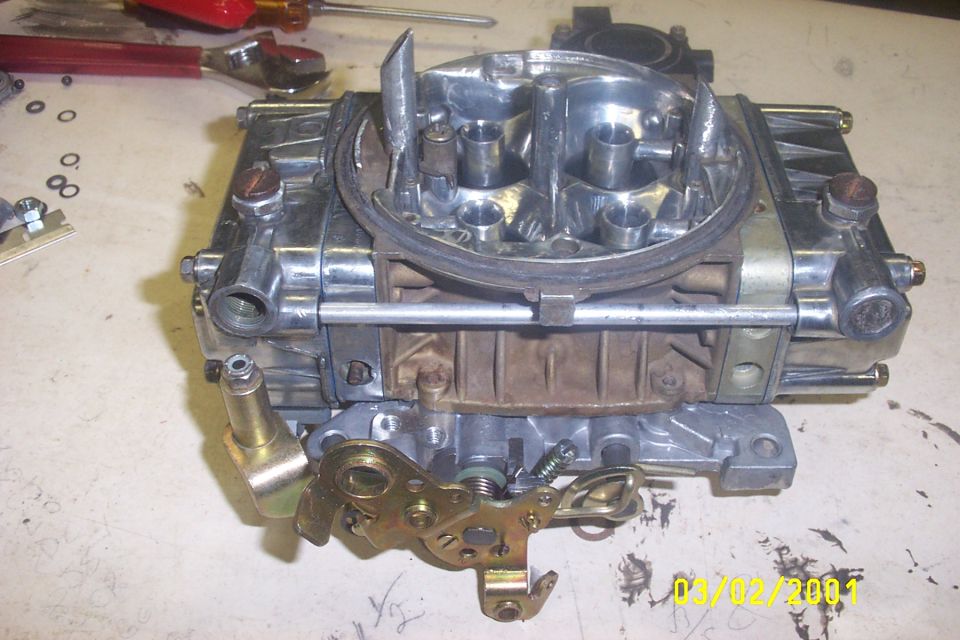 Carburetor with chrome accents and brass linkage, resting on a workbench with tools and small parts scattered around
