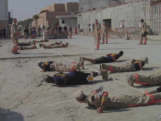 Service members doing push-ups on sand at a desert base while instructors watch
