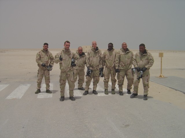 Group of seven people in tan desert uniforms posing on pavement in a desert