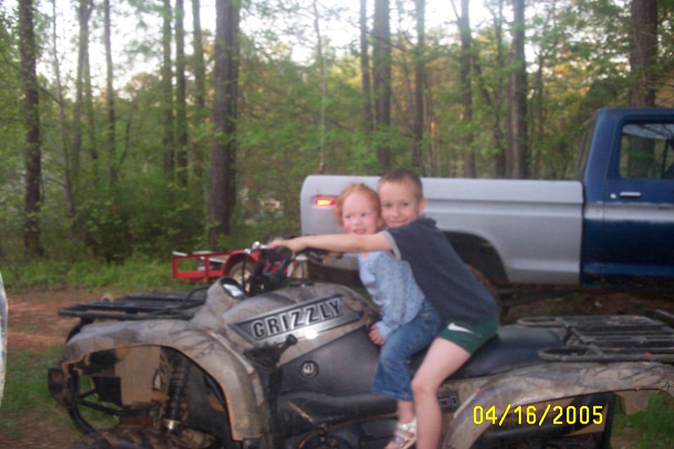 Two kids on a GRIZZLY ATV. A blue and silver pickup truck is behind them