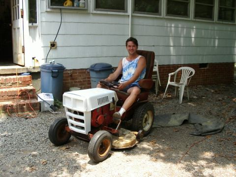 1985 Ford Bronco - A man sits on a riding lawn mower in front of a house with trash cans nearby.