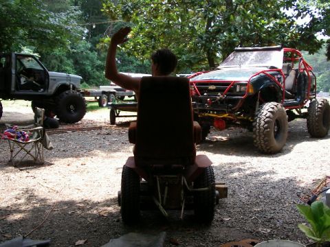 1985 Ford Bronco - A person on a riding mower is seen with two heavily modified off-road vehicles in a yard.