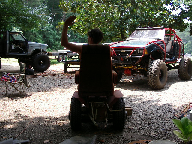 Person on riding mower waves, with a heavily modified off-road vehicle and a lifted truck in the background