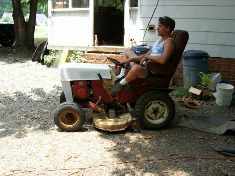 1985 Ford Bronco - A man sits on a riding lawn mower in a gravel driveway next to a house.