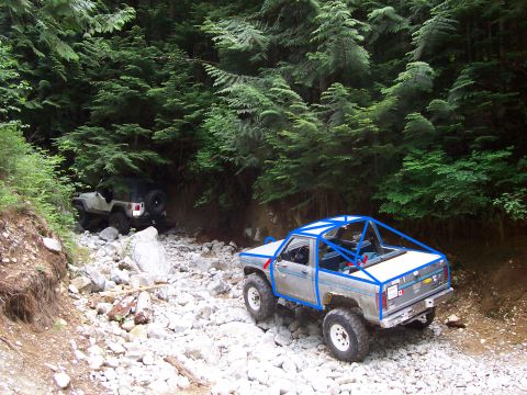 1984 Ford Bronco II - Two off-road vehicles navigate a rocky, wooded trail, with one truck prominently a blue roll cage.