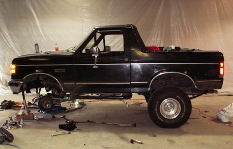 1988 Ford Bronco - Side view of a black full-size Bronco in a plastic-sheeted shop with its hardtop removed and the front fender, inner fender and front wheel…