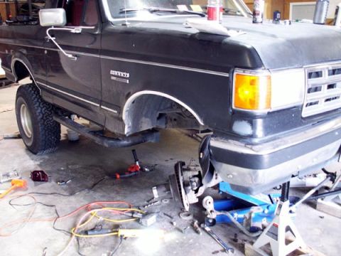 1988 Ford Bronco - A black full-size Ford Bronco sits in a garage with its right front wheel removed and the front end supported on a floor jack and jack stan…