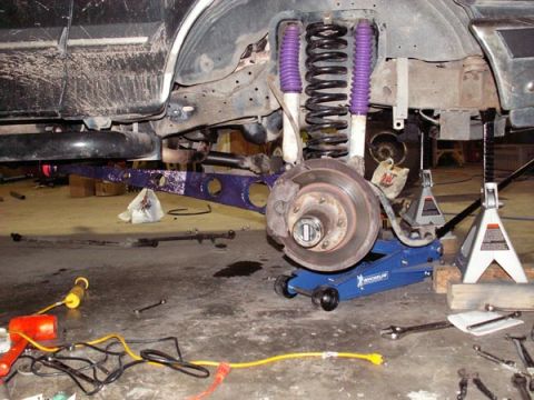 1988 Ford Bronco - Right front suspension of a lifted truck torn down in a garage, with a purple-powdercoated radius arm, black coil spring and two purple-boo…