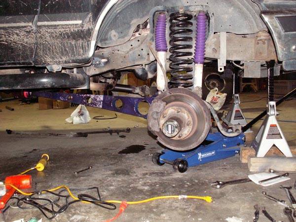 Front suspension teardown with purple radius arm, coil spring and bare brake rotor on floor jack