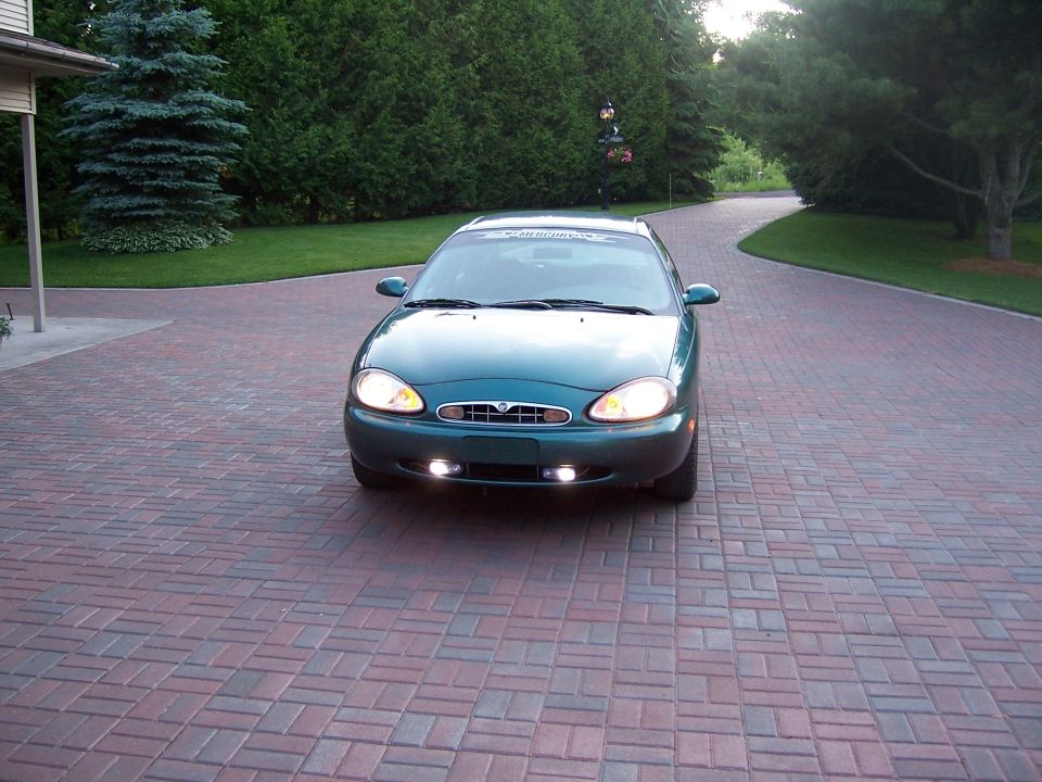 Green Mercury Sable sedan front view with headlights and fog lights on in a paver driveway