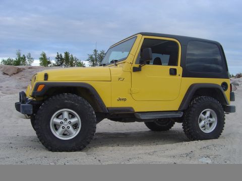 1986 Ford F-150 - A yellow Jeep Wrangler TJ is parked on a sandy surface with a rocky, tree-lined background.