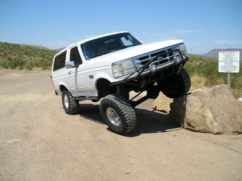 1993 Ford Bronco - White Ford Bronco with lifted suspension and off-road tires climbing a rock on a dirt path.