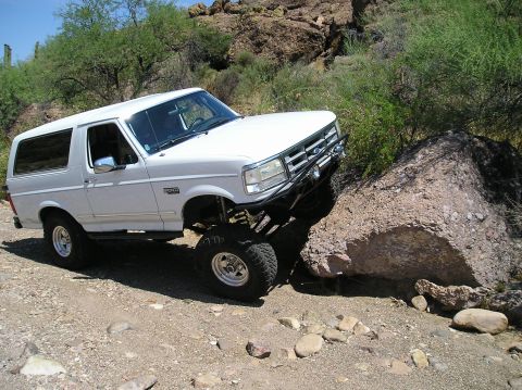 1993 Ford Bronco - White Ford Bronco with lifted suspension is shown off-roading on a rocky, desert-like trail.
