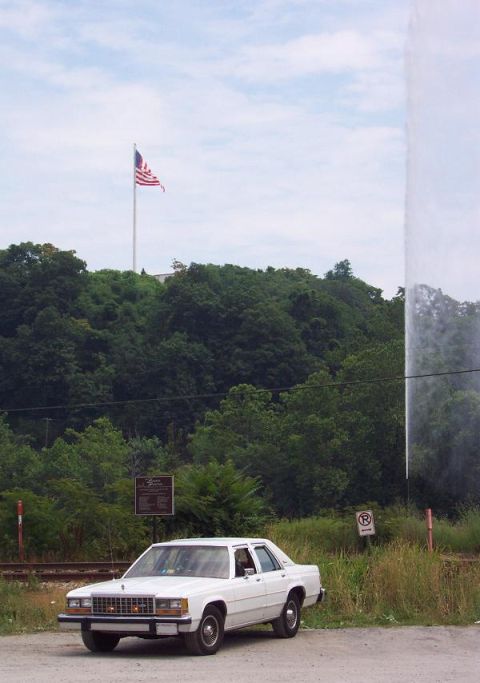 1985 Ford Crown Victoria LTD - A white sedan is parked on a dirt road, with a flagpole and trees in the background.