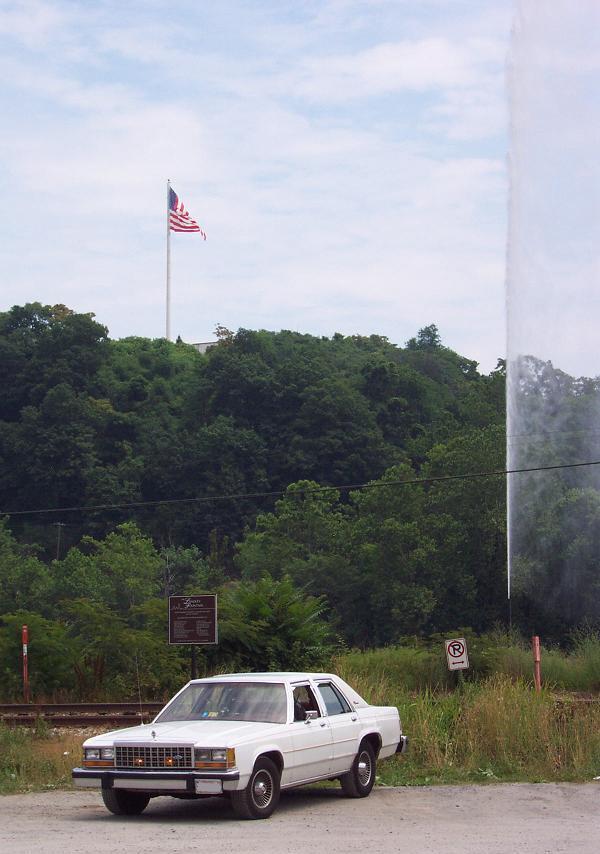 White sedan parked on a dirt road with trees and a flagpole in the background
