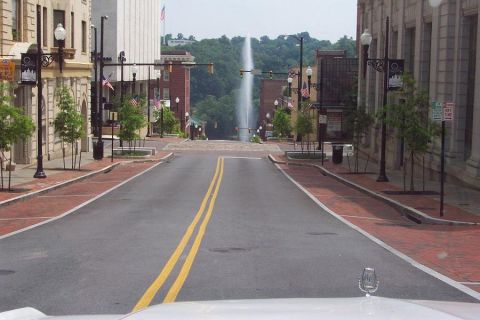 1985 Ford Crown Victoria LTD - A street scene with a prominent fountain in the distance, flanked by buildings and trees.