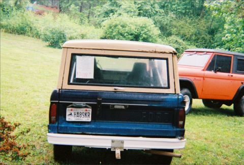 1967 Ford Bronco - Two Ford Broncos are parked in a grassy area with trees in the background. One is blue and the other is orange.
