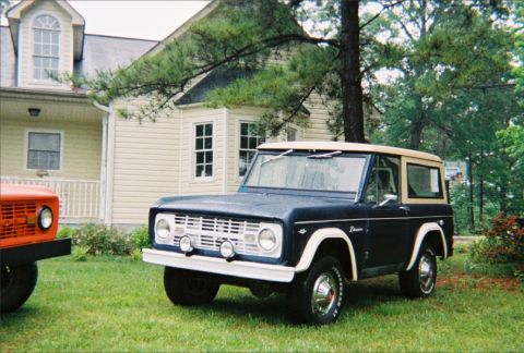 1967 Ford Bronco - A dark blue Ford Bronco with a tan top is parked on a grassy lawn in front of a house, with another vehicle visible to the left.